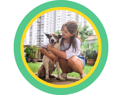 A woman outside in front of a white building hugging a dog in the grass